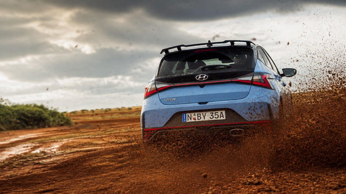 Blauer Hyundai i20 N fährt schnell auf matschiger Offroad-Strecke, schlamm spritzt nach hinten.