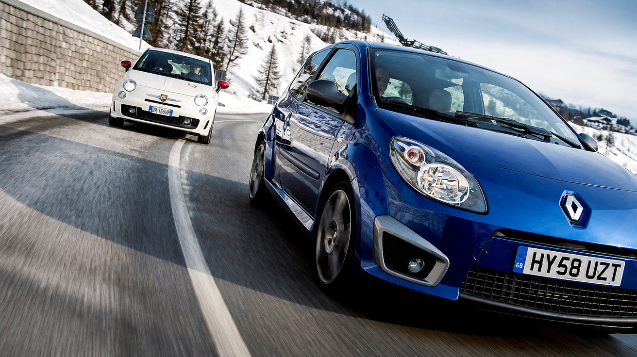 Blauer Renault und weißer Fiat fahren auf kurviger Bergstraße mit Schneelandschaft im Hintergrund.