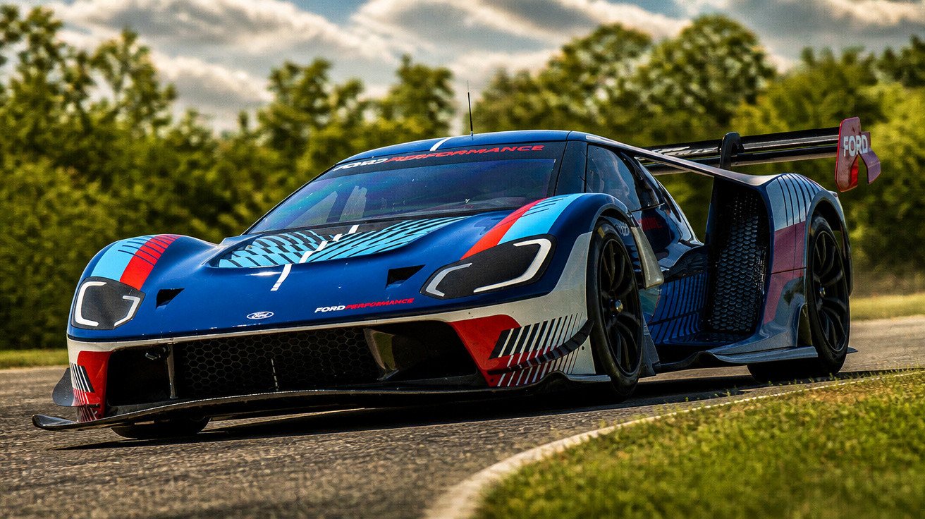 Blau-rotes Ford GT Rennfahrzeug auf Rennstrecke mit Bäumen im Hintergrund bei Sonnenlicht.