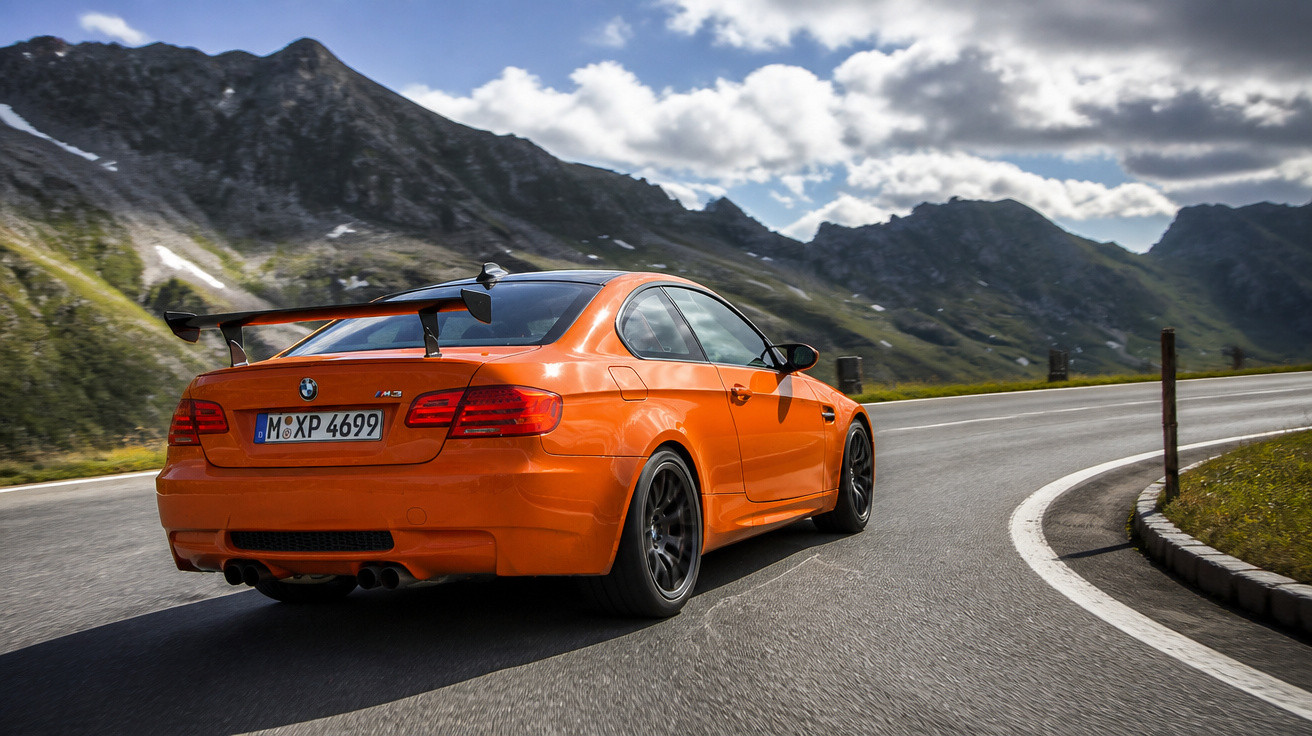 Oranger BMW M3 fährt auf kurviger Bergstraße mit bewölktem Himmel und Bergpanorama im Hintergrund.