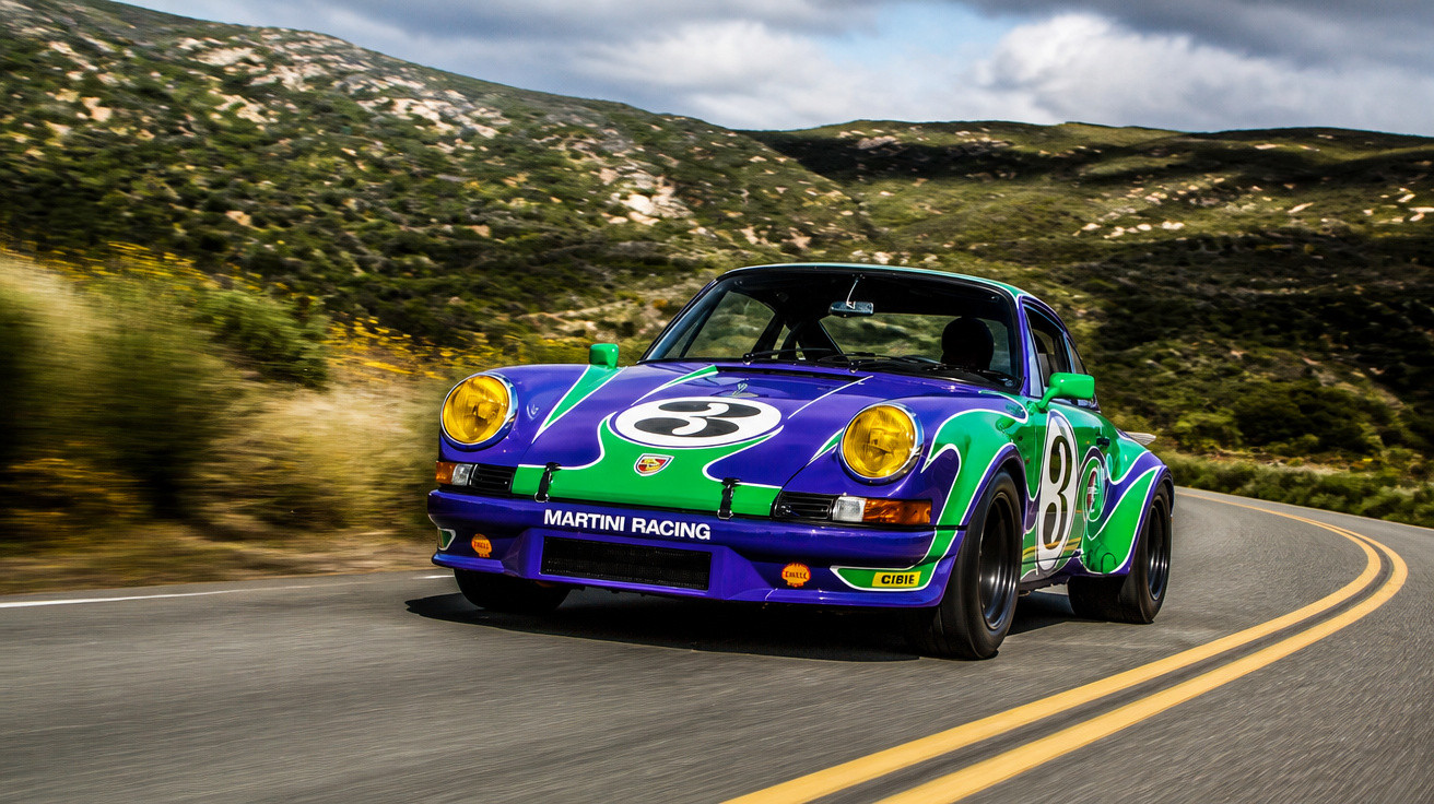 Blau-grüner Porsche im Martini Racing Design fährt auf kurviger Bergstraße bei bewölktem Himmel.