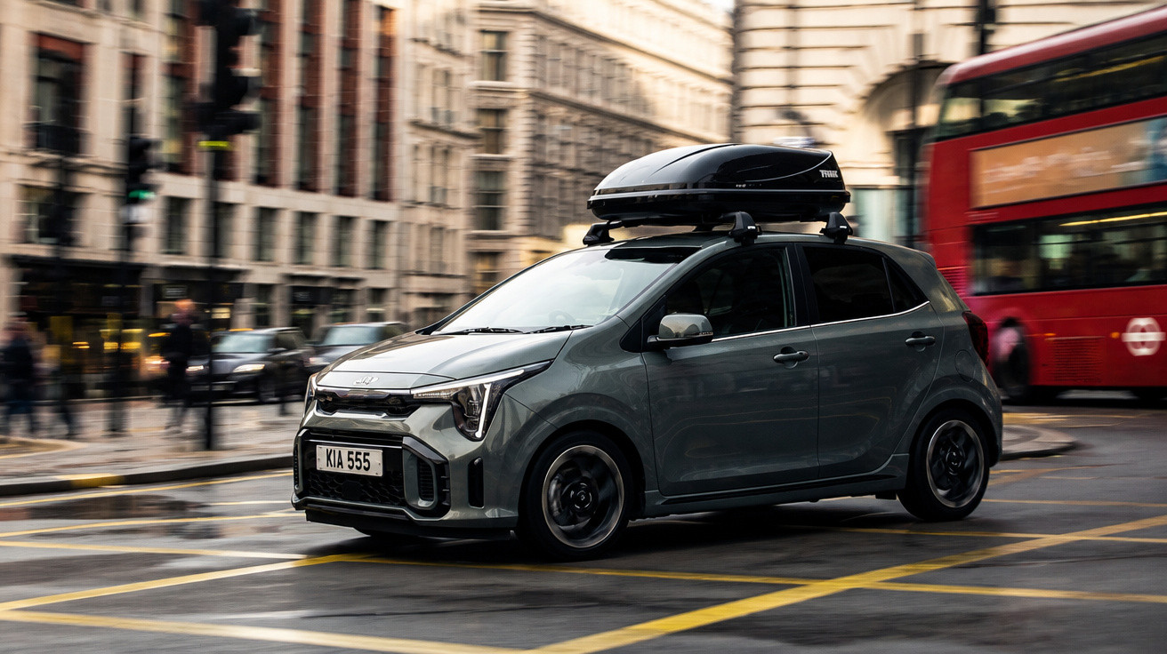 Kleiner grauer Kia mit Dachbox fährt in Stadt mit rotem Doppeldeckerbus im Hintergrund.