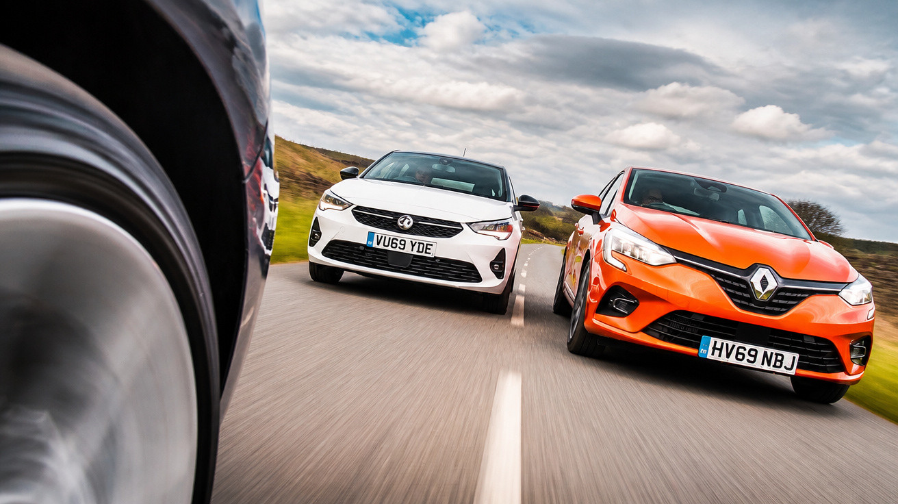 Zwei moderne Autos, ein weißer Opel und ein orangefarbener Renault, fahren nebeneinander auf einer Landstraße.