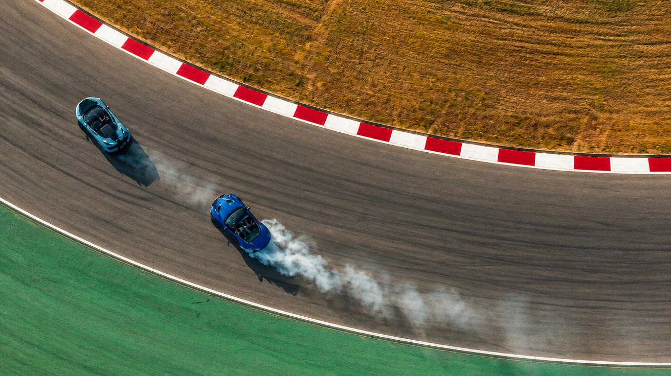 Zwei blaue Sportwagen driftet auf einer Rennstrecke in einer Kurve, Rauch hinter einem Auto sichtbar.