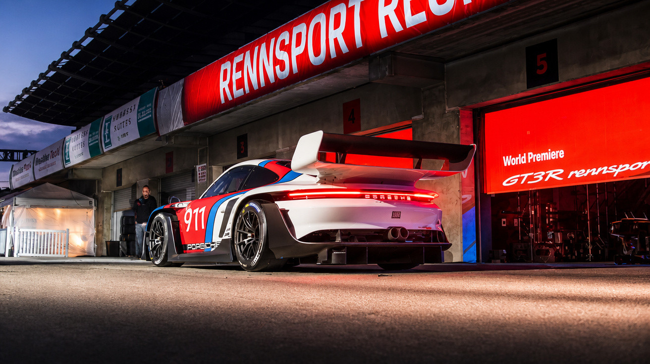 Rennsport-Porsche 911 mit großer Heckflosse vor Boxencrew und rotem Rennsport-Logo bei Abendlicht.