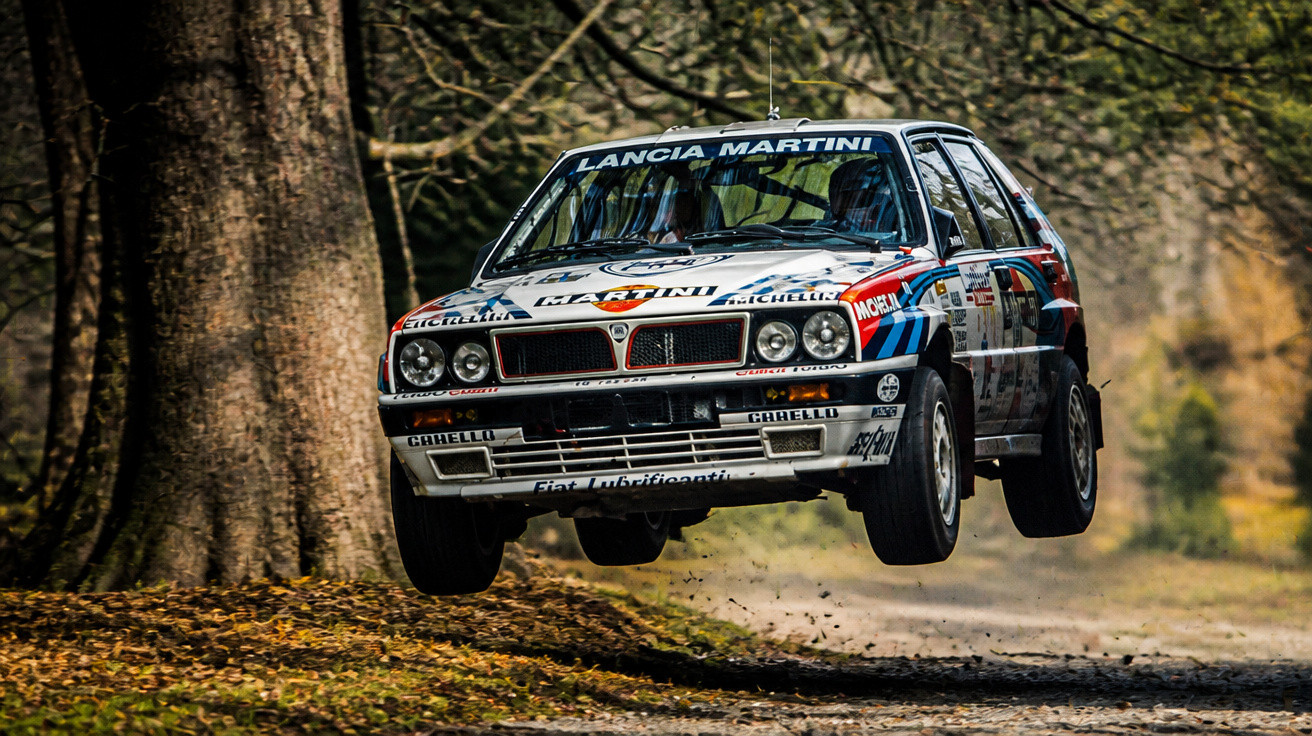 Lancia Martini Rallyeauto springt auf einer Waldstrecke mit Laub auf dem Boden.