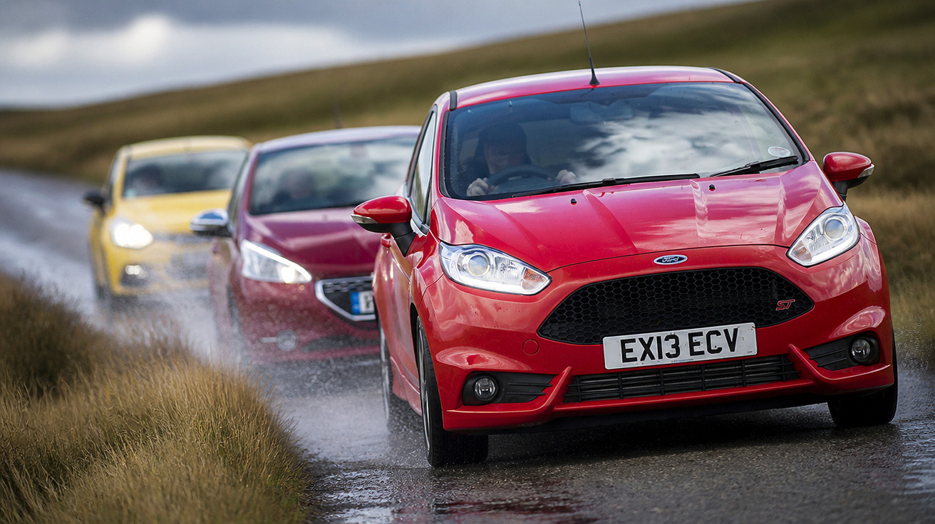Drei Autos fahren hintereinander auf einer nassen Landstraße, vorne ein roter Ford Fiesta.