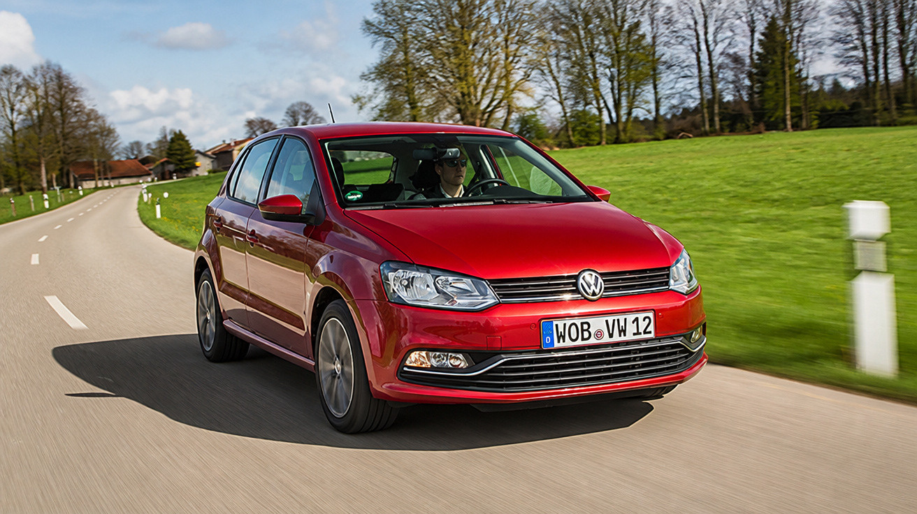Roter VW Polo fährt auf Landstraße mit grünem Gras und Bäumen bei Tageslicht.