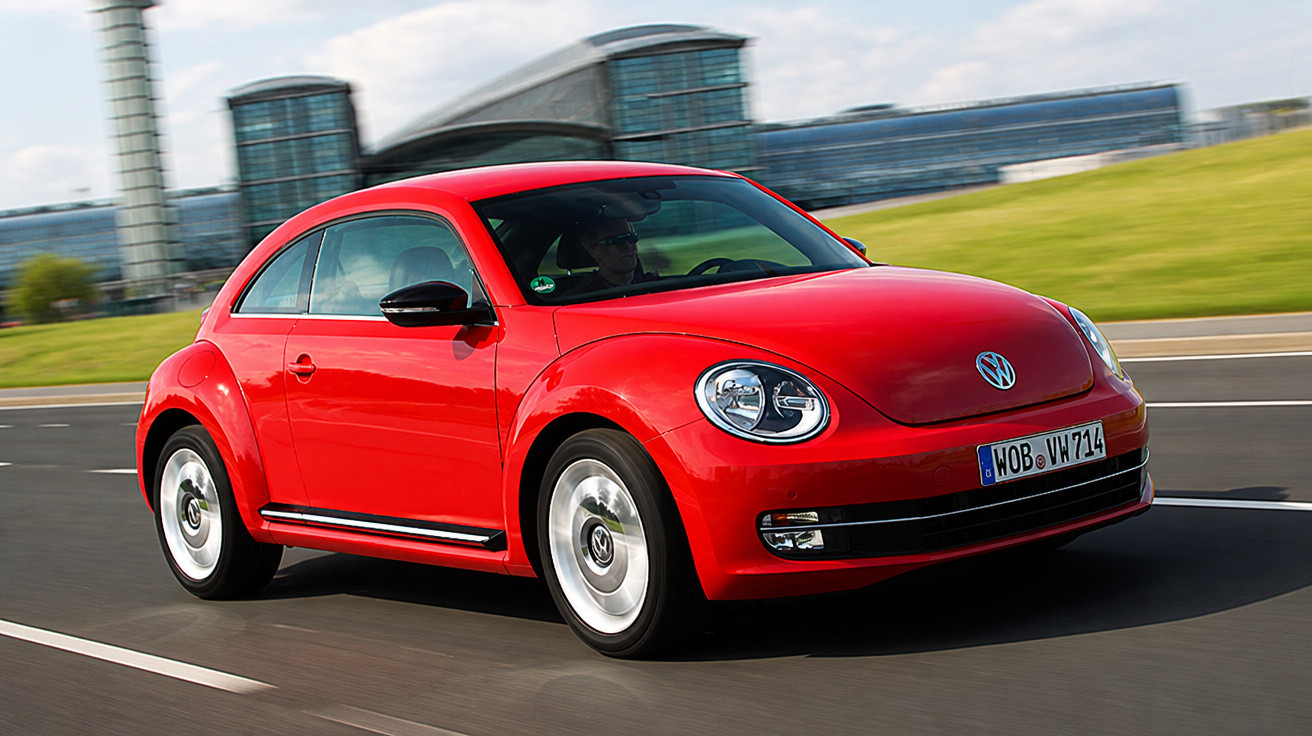 Roter Volkswagen Beetle fährt auf mehrspuriger Straße mit moderner Glasarchitektur im Hintergrund.